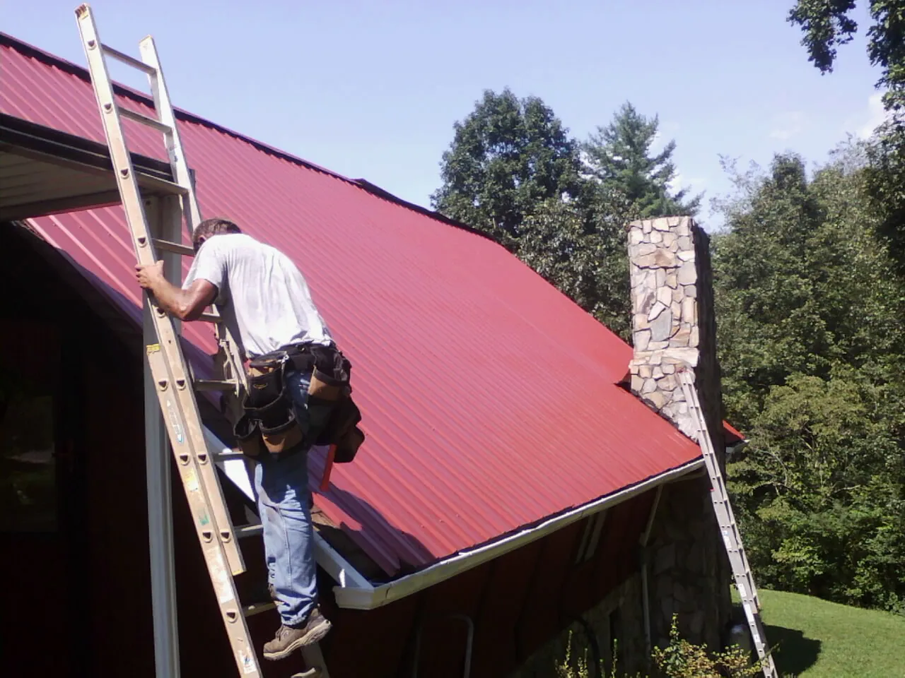 Roofer on a ladder beside a red metal roof and stone chimney — Soffit & Fascia Repair by Roofing Brothers in Carthage
